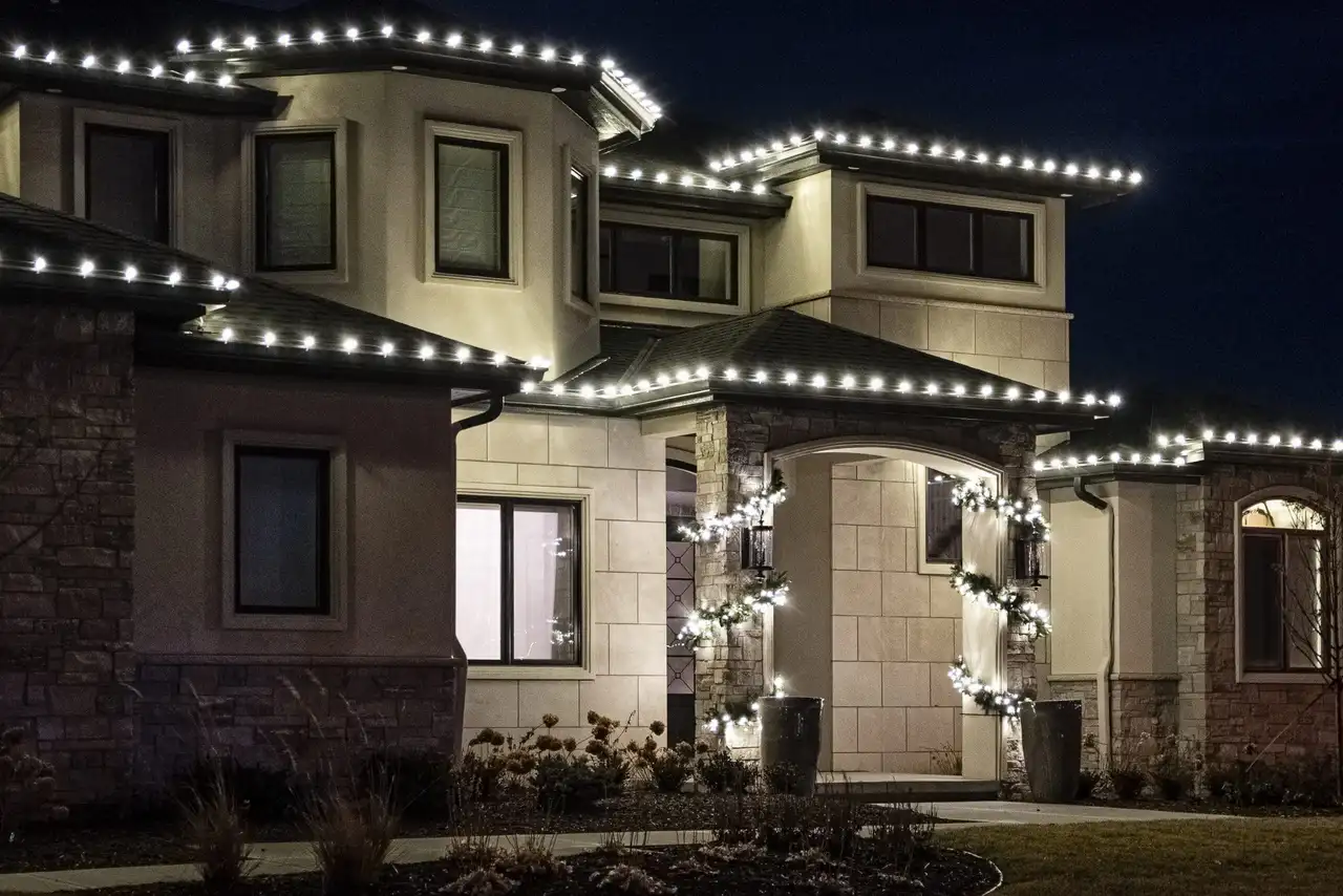Two-story home at night with cool white Christmas lights along the rooflines and lit garland on the entry columns