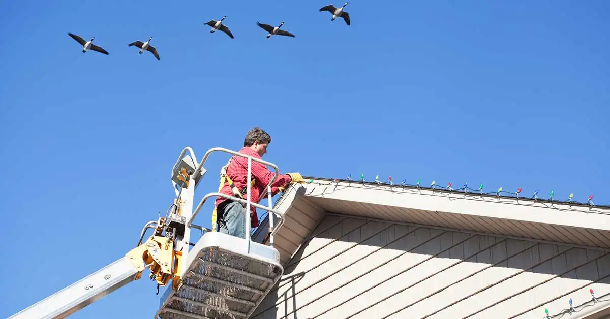 Installer in a bucket lift attaching multicolor Christmas lights along a roofline under a blue sky